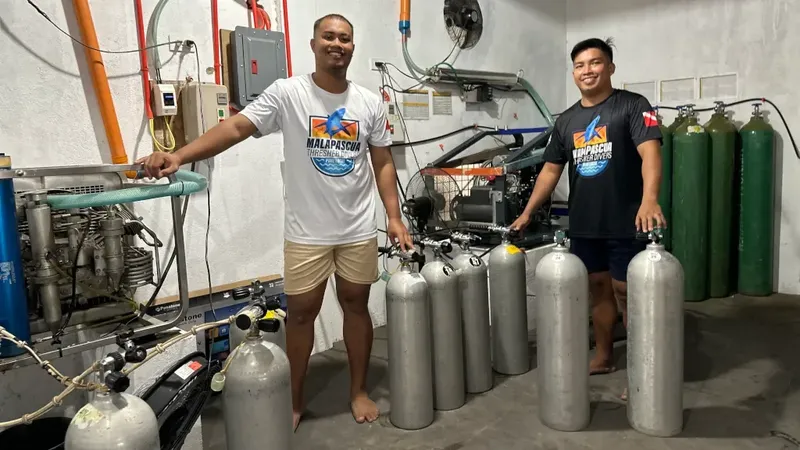 Deux hommes souriants posent devant des bouteilles de plongée et du matériel