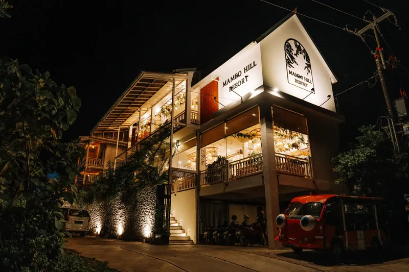 Bâtiment éclairé la nuit avec enseigne Mambo Hill Resort et véhicules