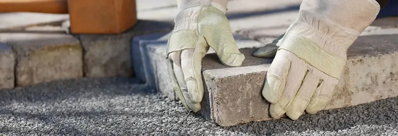 Mains gantées posant des pavés sur un lit de gravier clair