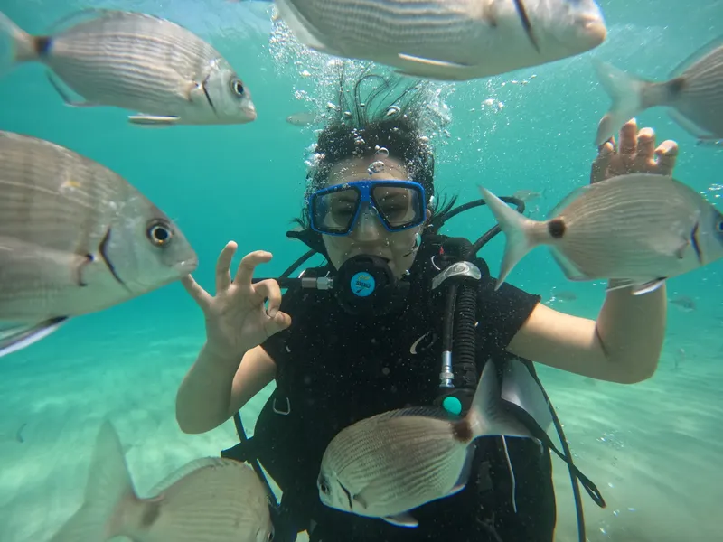 Plongeuse souriante entourée de nombreux poissons argentés dans l'eau turquoise