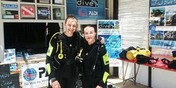 Deux plongeuses souriantes en combinaison devant un centre de plongée