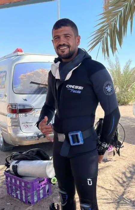 Plongeur souriant en combinaison, pouce levé, devant un van et du matériel