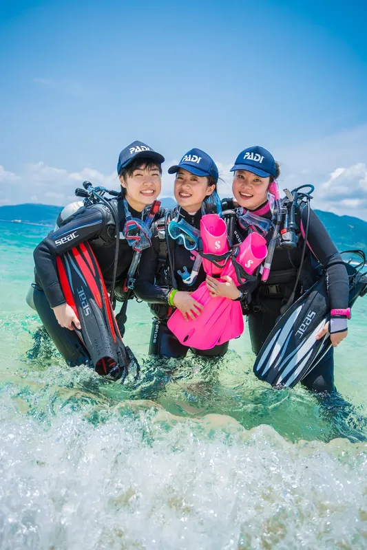 Trois plongeuses souriantes équipées sur une plage ensoleillée