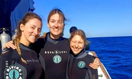 Trois plongeuses souriantes sur un bateau en mer, équipement visible