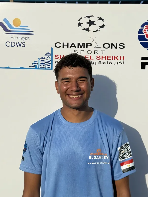 Un homme souriant, portant un t-shirt bleu, devant des bannières