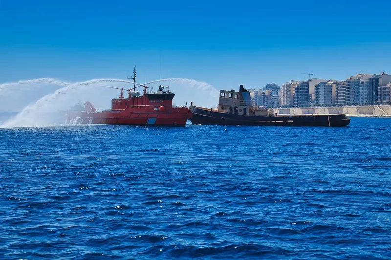 Deux bateaux en mer, un rouge lance un jet d'eau, ville en fond