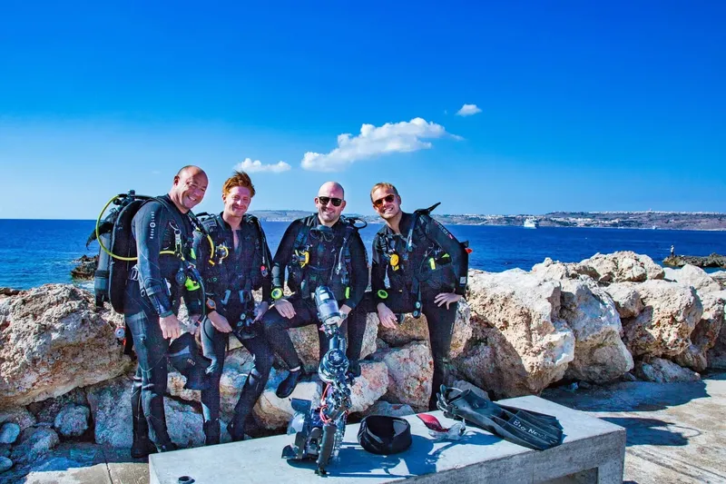 Quatre plongeurs souriants en combinaison sur un quai rocheux ensoleillé