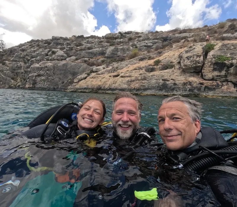 Trois plongeurs souriants, équipés, émergent dans une eau claire et turquoise
