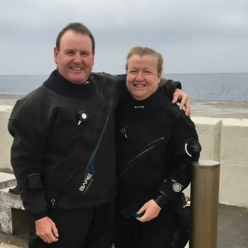 Deux plongeurs souriants en combinaison, bras dessus bras dessous, au bord de l'eau