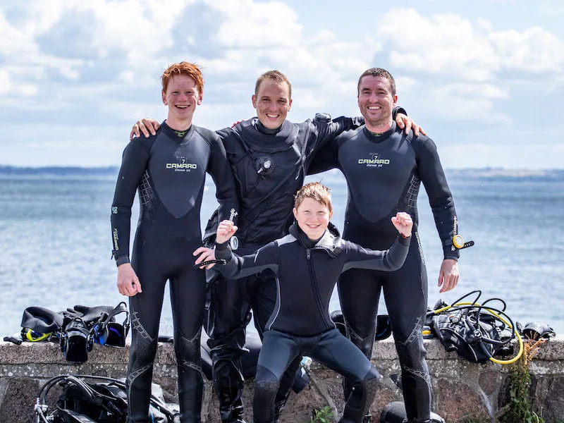 Quatre plongeurs souriants en combinaison, devant la mer et leur équipement