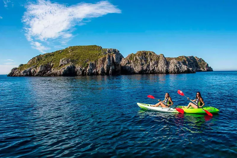 Deux femmes pagayent en kayak sur une mer bleue face à une île rocheuse