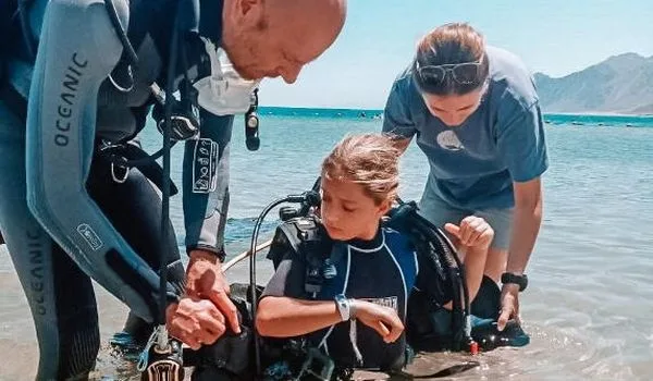 Trois personnes équipent un jeune plongeur sur une plage ensoleillée