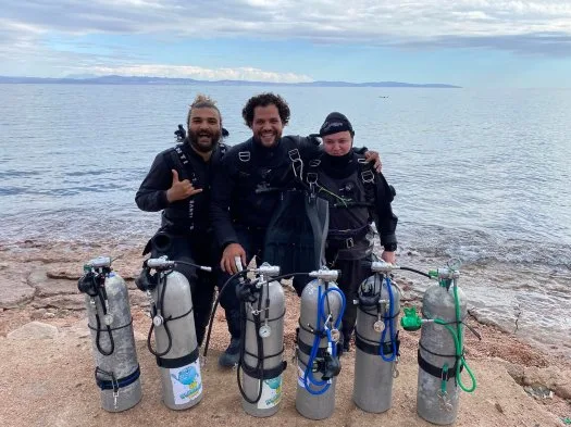 Trois plongeurs souriants posent avec leur équipement sur un rivage rocheux