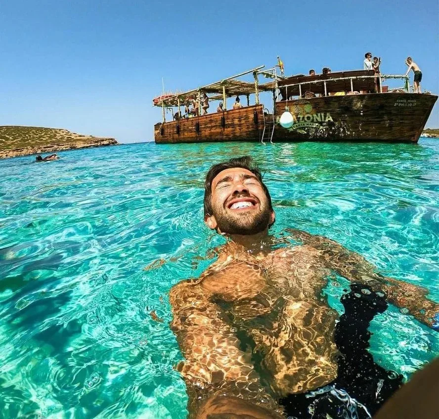 Homme souriant dans une eau turquoise avec un bateau en arrière-plan