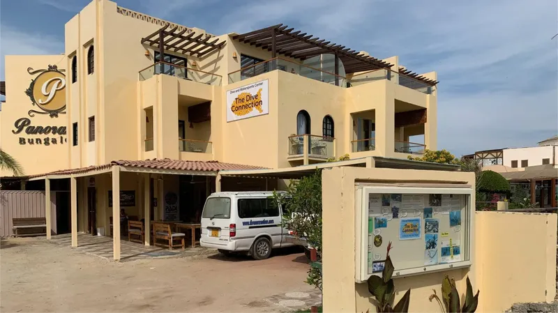 Bâtiment jaune avec pergola, van blanc, panneau d'affichage et végétation