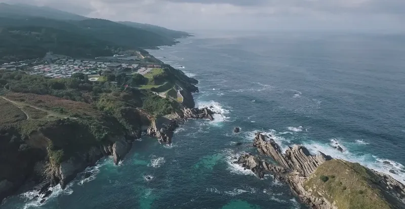 Côte rocheuse avec végétation, bâtiments et mer sous ciel nuageux