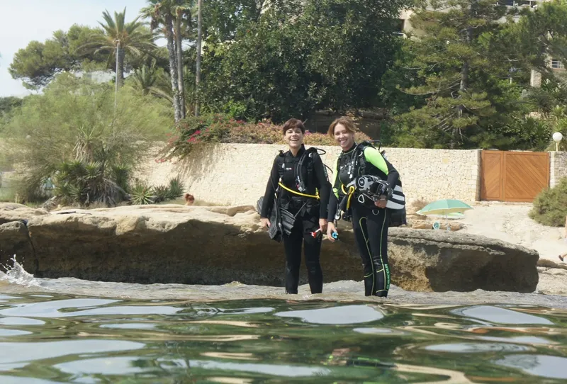 Deux plongeuses souriantes équipées sur une plage rocheuse ensoleillée
