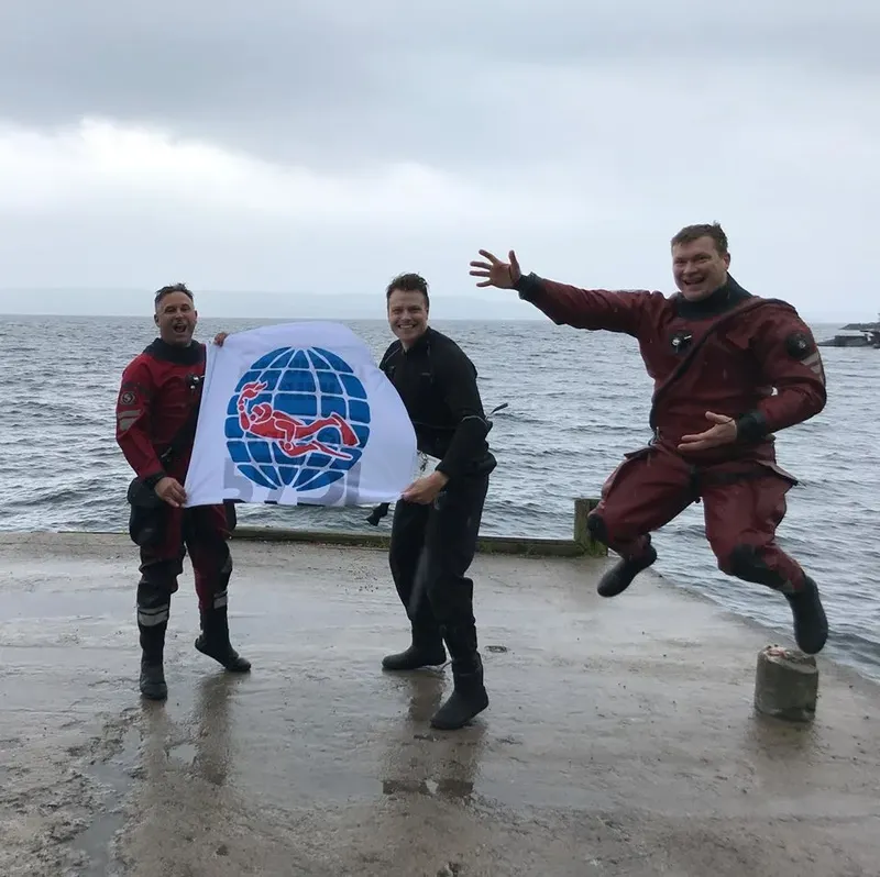 Trois plongeurs souriants posent sur un quai gris face à la mer