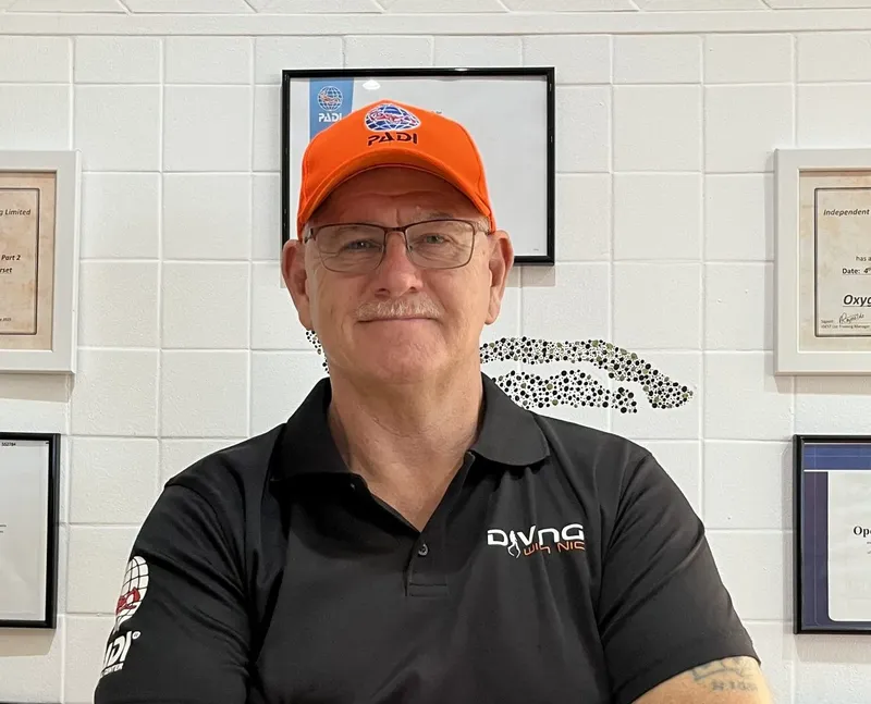 Homme souriant portant un polo et une casquette orange devant des certificats