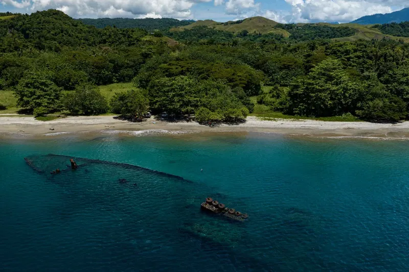 Epave de bateau visible sous l'eau turquoise, plage et végétation luxuriante