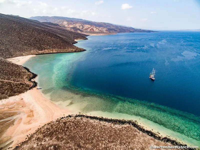 Vue aérienne d'une côte rocheuse, mer turquoise et voilier au loin