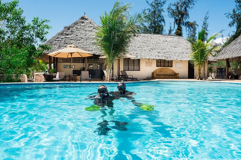 Deux plongeurs s'entrainent dans une piscine devant un bâtiment tropical
