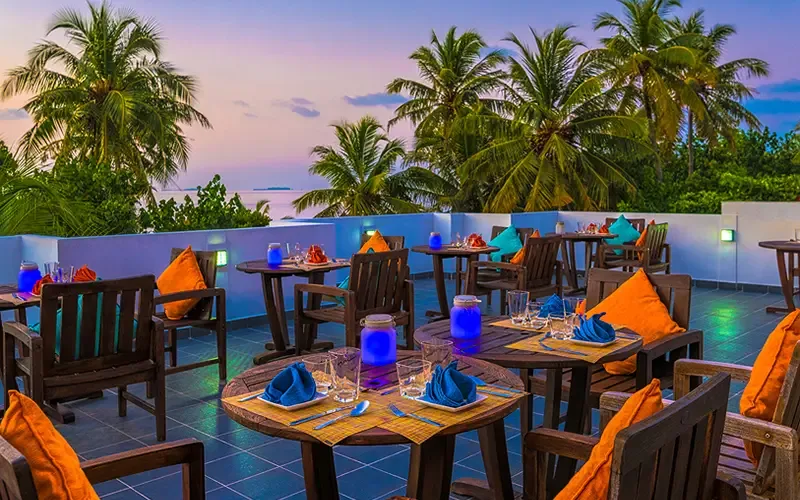 Terrasse restaurant avec tables préparées, palmiers et ciel coloré