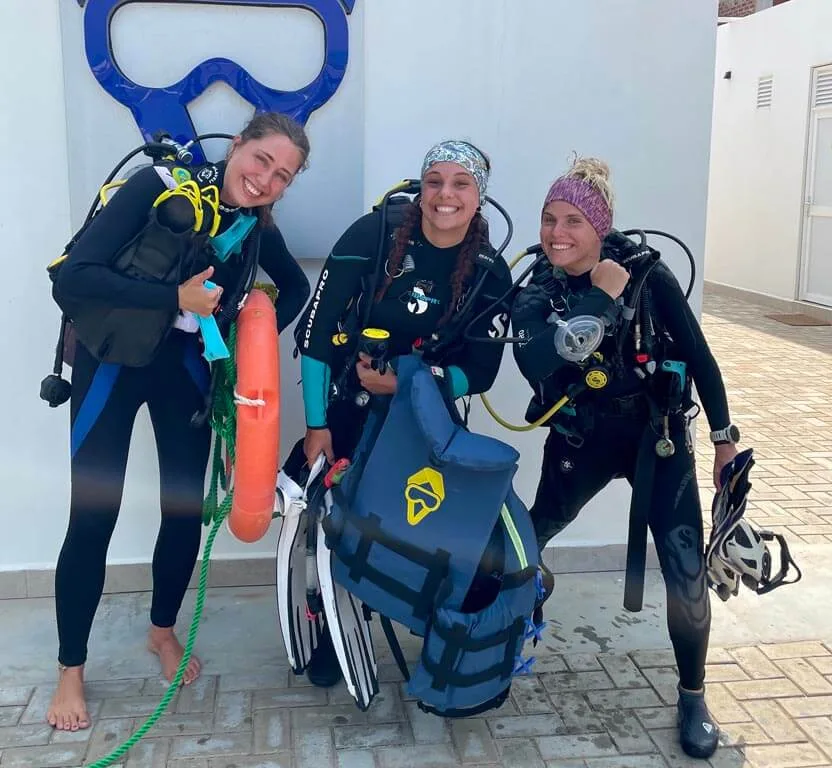 Trois plongeuses souriantes équipées près d'un mur blanc ensoleillé