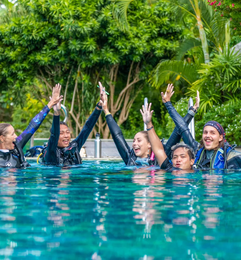 Groupe de plongeurs souriants, bras levés, dans une piscine turquoise