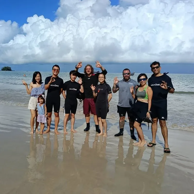 Groupe de personnes souriantes sur une plage ensoleillée, vêtues de tenues de plongée