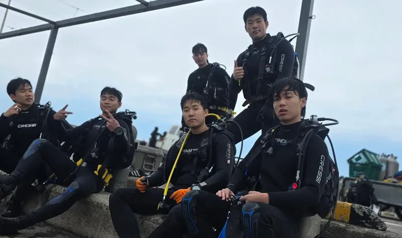 Groupe de plongeurs équipés sur un quai, souriant et faisant des signes