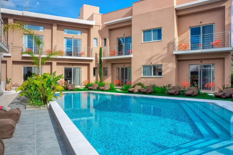 Une piscine turquoise au premier plan, devant un bâtiment beige à plusieurs étages avec des balcons.