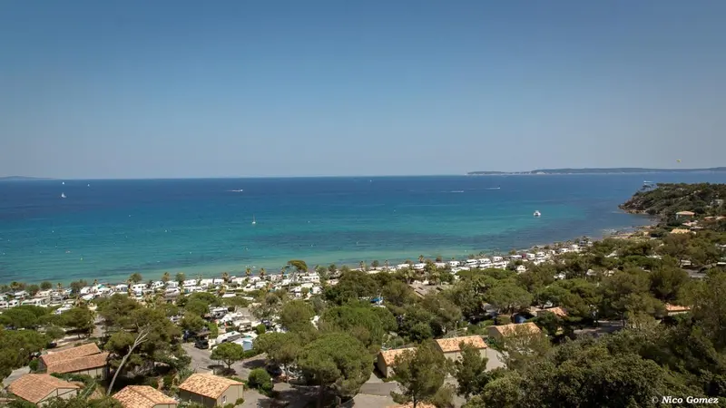 Vue aérienne d'une plage bordée de pins et de bâtiments bas.