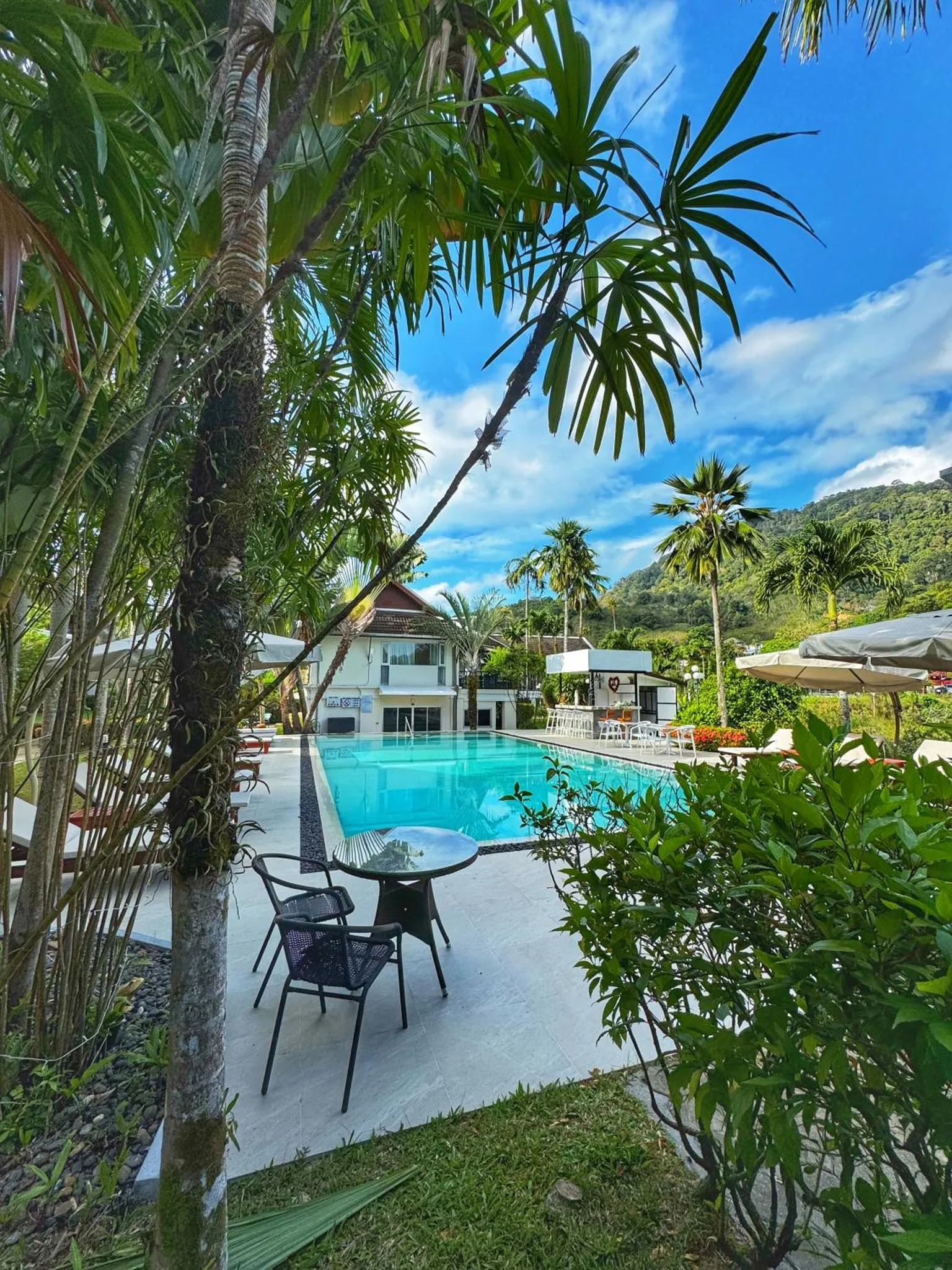 Une table et des chaises sont placées près d'une piscine ensoleillée entourée de végétation tropicale.