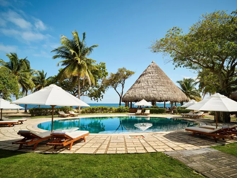 Une piscine bordée de chaises longues sous des parasols, avec une structure de toit de chaume en arrière-plan.