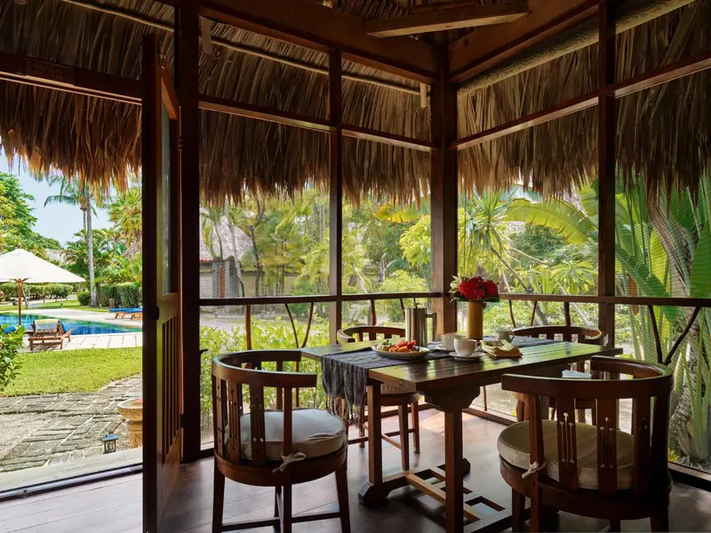 Une table et des chaises en bois sont disposées sur une terrasse ombragée avec vue sur un jardin et une piscine.