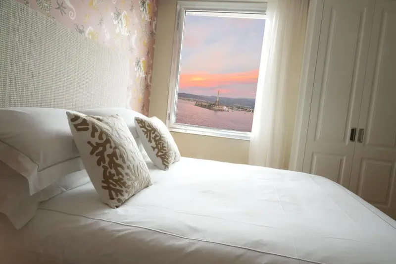 Un lit avec des oreillers face à une fenêtre donnant sur un paysage côtier au coucher du soleil.