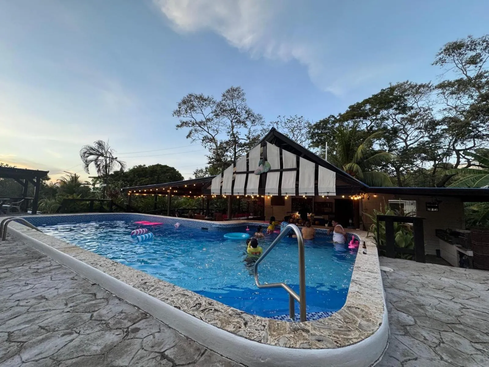 Une piscine rectangulaire bordée d'une terrasse en pierre, avec des personnes se baignant et un bâtiment à l'arrière-plan.
