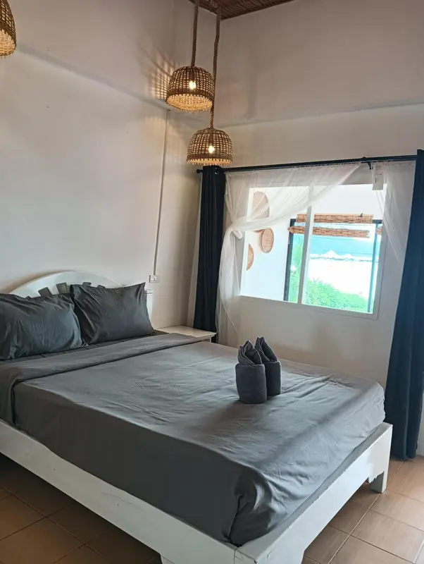 Une chambre lumineuse avec un lit recouvert d'un drapé gris et une fenêtre ouverte sur la végétation.