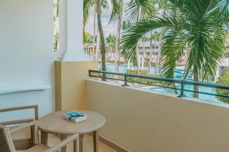 Une terrasse avec une table et des chaises offre une vue sur une piscine et une végétation luxuriante.