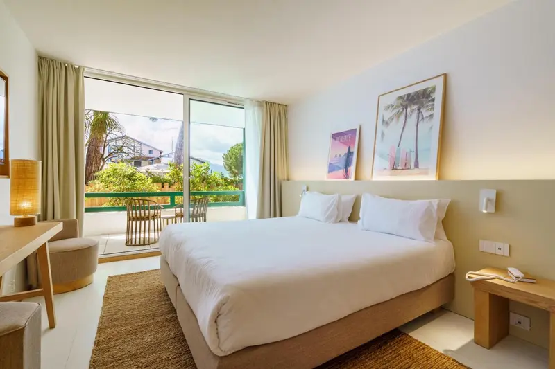 Chambre lumineuse avec un grand lit et une vue sur une terrasse et la végétation.