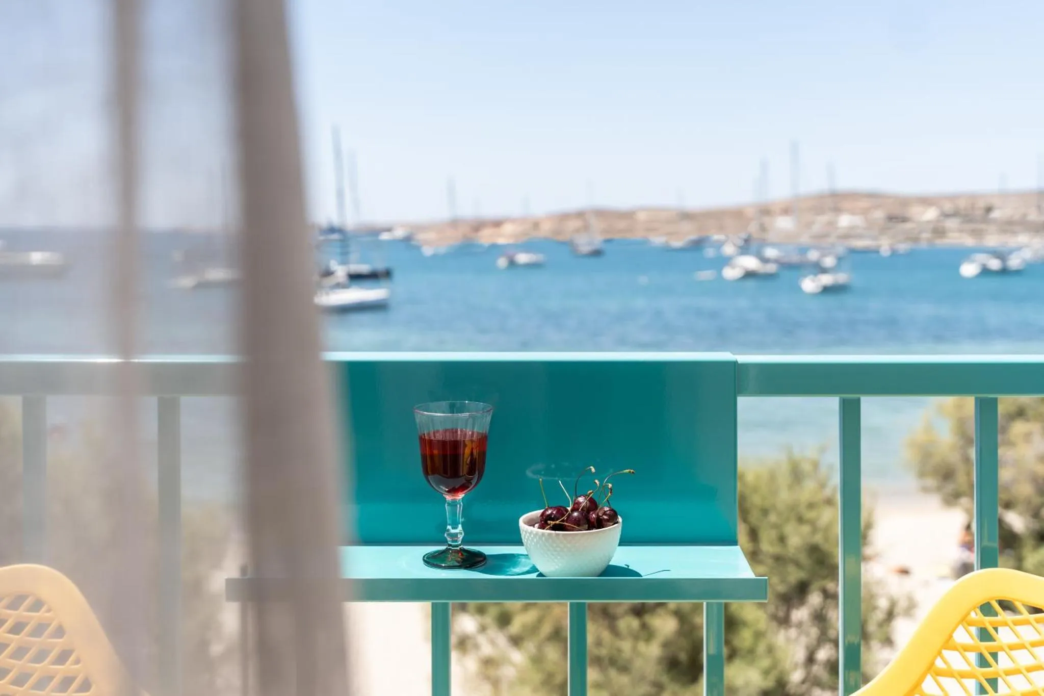Une table avec des boissons et des fruits est posée sur une terrasse ensoleillée avec vue sur la mer et des bateaux.