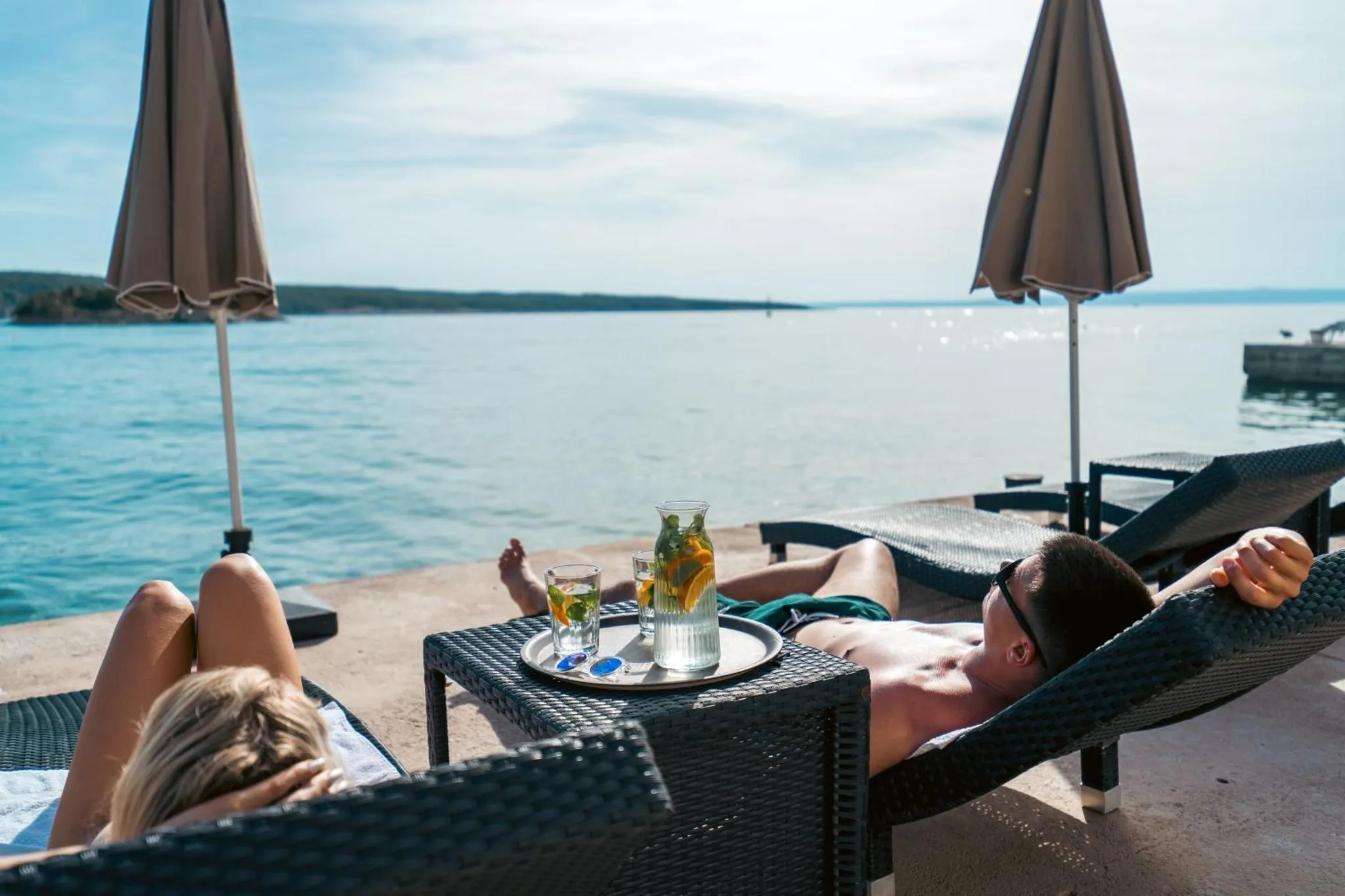 Deux personnes se relaxent sur des transats au bord de l'eau avec des boissons.