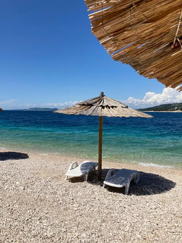 Deux transats blancs sur une plage de galets sous des parasols.