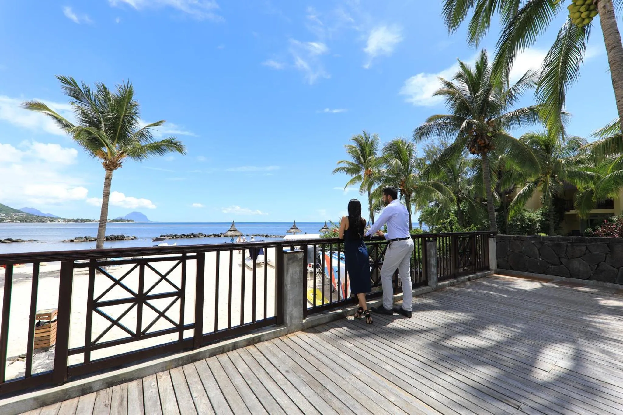 Deux personnes se tiennent sur une terrasse en bois face à la mer et à la végétation tropicale.