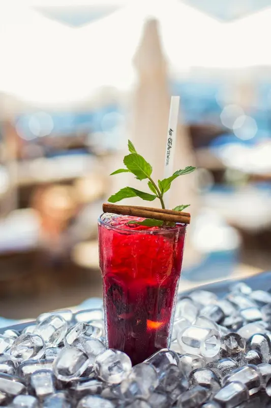 Un verre de boisson rouge orné de menthe et d'une cannelle, posé sur une surface réfléchissante.