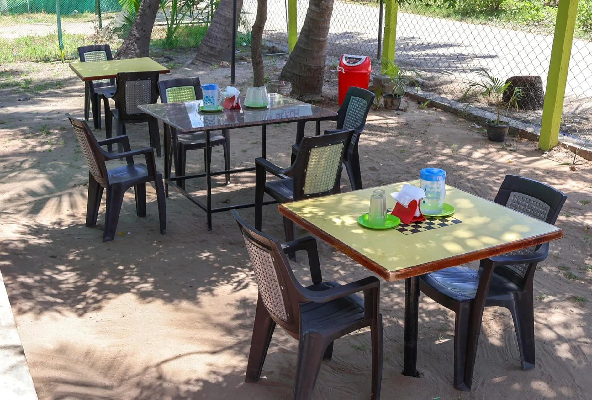 Tables et chaises en plastique disposées sur un sol sablonneux à l'ombre de palmiers.