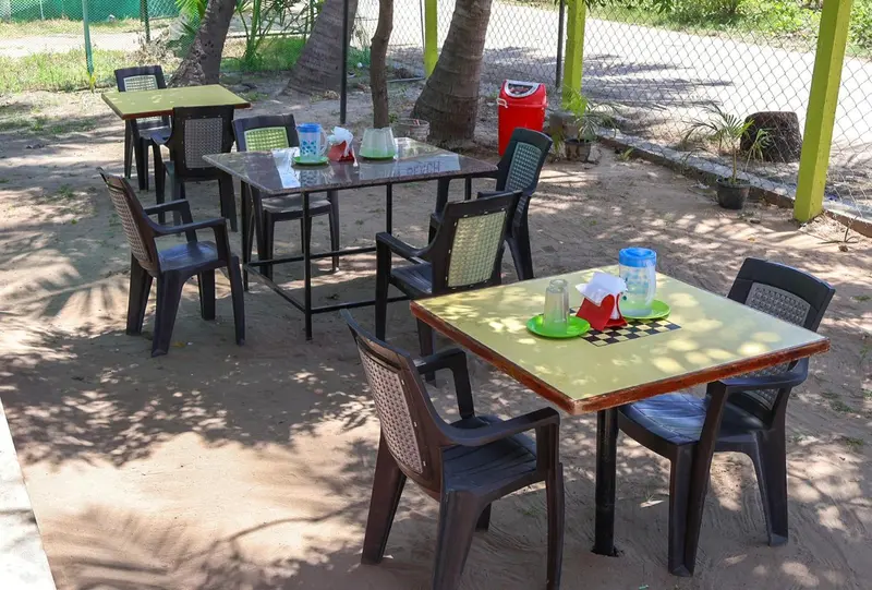 Tables et chaises en plastique disposées sur un sol sablonneux à l'ombre de palmiers.