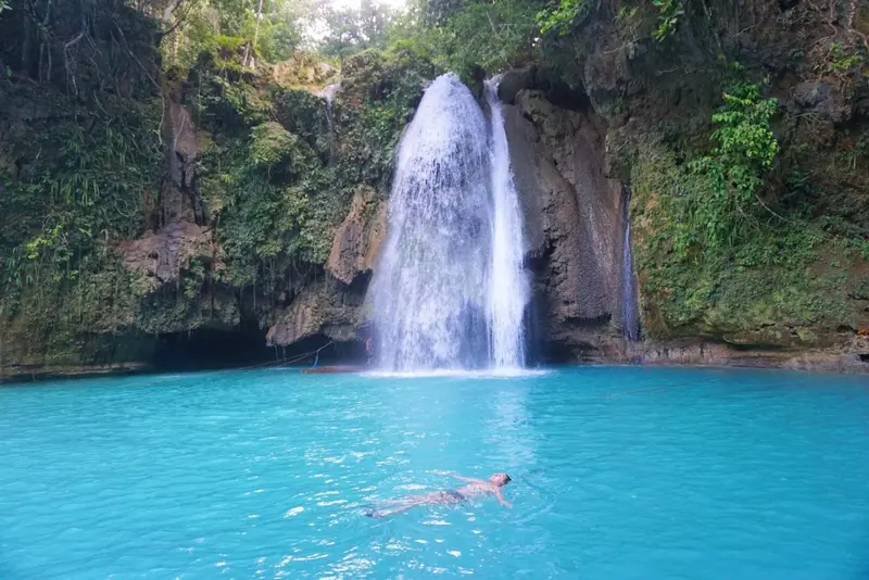 Une cascade se jette dans une piscine naturelle turquoise où une personne nage sur le dos.