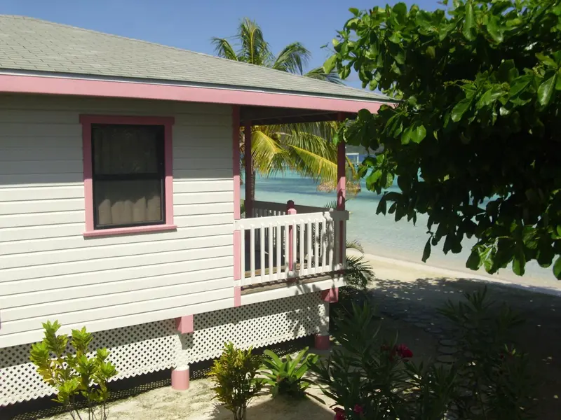 Petite maison blanche avec des volets roses et une terrasse en bois donnant sur une plage.